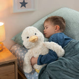 A young boy in blue pajamas sleeps soundly in bed, hugging a large white fluffy duck plush toy. The scene is set in a cozy bedroom with a lit bedside lamp, patterned bedding, and a mint green pillow.