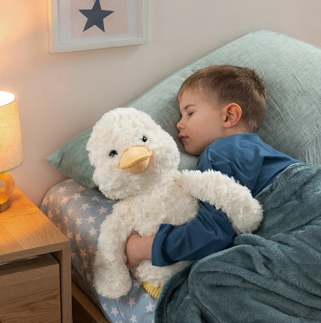 A young boy in blue pajamas sleeps soundly in bed, hugging a large white fluffy duck plush toy. The scene is set in a cozy bedroom with a lit bedside lamp, patterned bedding, and a mint green pillow.