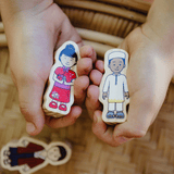 Two small wooden toy figures with painted cartoon children are held in a person's hands over a woven brown surface. One figure wears a red outfit and the other wears a white outfit.