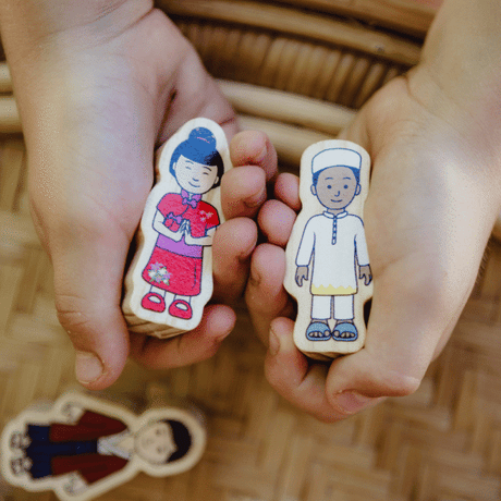 Two small wooden toy figures with painted cartoon children are held in a person's hands over a woven brown surface. One figure wears a red outfit and the other wears a white outfit.