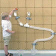 A young child in a white shirt and khaki shorts stands beside a wall-mounted transparent plastic water pipe toy, pressing a small orange button to release water into clear measuring cups on the floor.