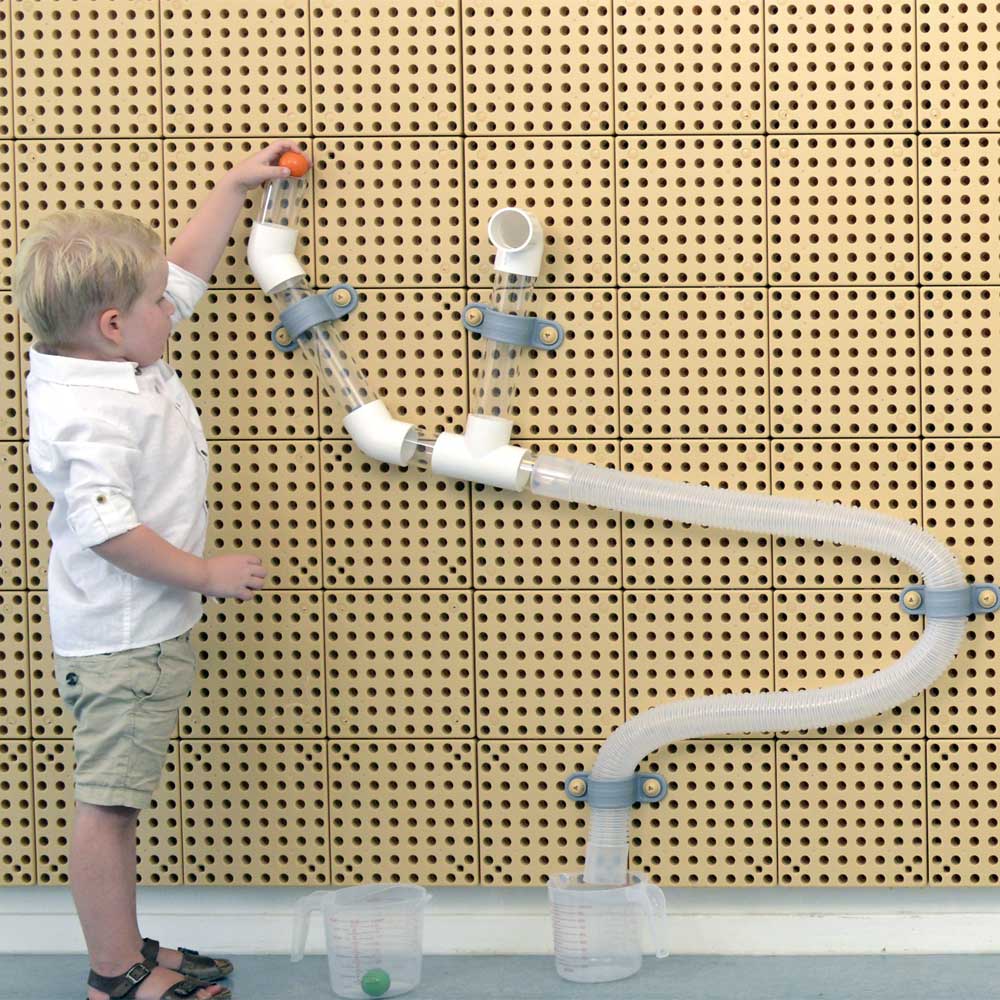 A young child in a white shirt and khaki shorts stands beside a wall-mounted transparent plastic water pipe toy, pressing a small orange button to release water into clear measuring cups on the floor.