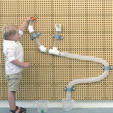 A young child in a white shirt and khaki shorts stands beside a wall-mounted transparent plastic water pipe toy, pressing a small orange button to release water into clear measuring cups on the floor.