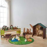 A children's farm-themed play set arranged in a bright room with a window and light wood floor. The set includes a green and brown wooden chicken coop, a wooden cart with green side panels, a round green farm rug, and several farm animal toys including chickens, ducks, a pig, and a cow.